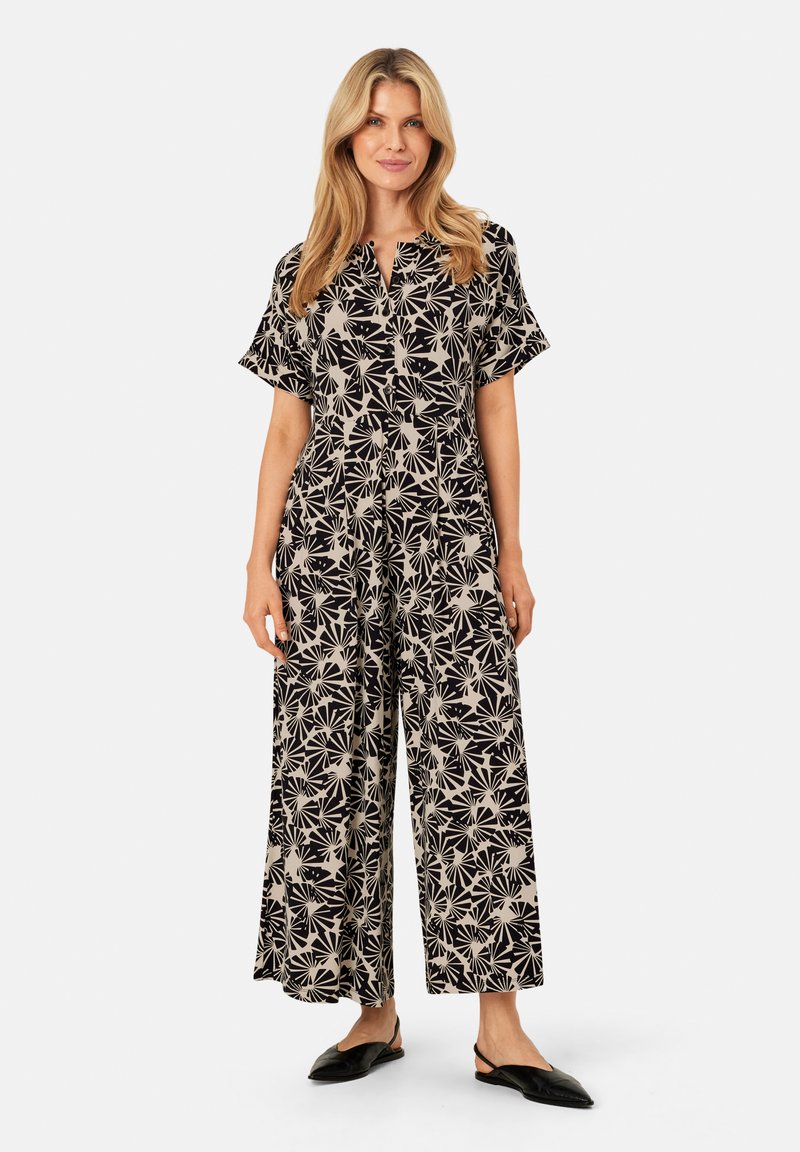 Schwarzer und beiger gemusterter Jumpsuit mit kurzen Ärmeln, Knopfverschluss, weitem Bein und Blumenmuster. Kombiniert mit schwarzen Slippers.