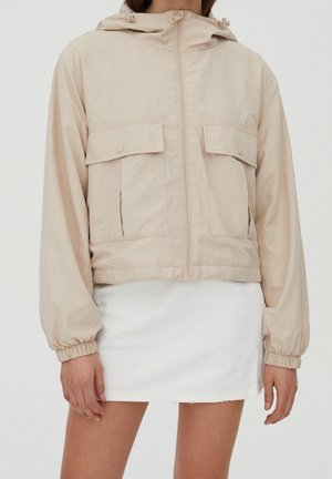 Personne portant une veste beige à capuche avec poches avant et poignets élastiques, associée à une mini-jupe blanche, sur un fond neutre.