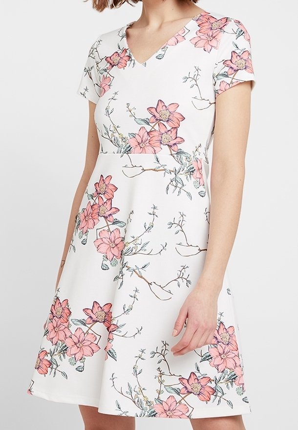 Robe blanche à manches courtes avec imprimé floral rose et encolure en V portée par une personne aux cheveux mi-longs.