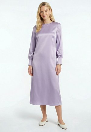 Femme portant une robe midi en satin violet clair à manches longues avec des chaussures blanches à petits talons, debout devant un fond blanc uni.