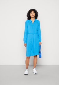 Vestito lungo a maniche lunghe di un blu brillante con l'apertura a bottoni e vita legata. La modella porta anche una piccola borsa blu e indossa delle sneakers bianche.