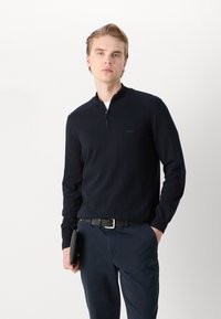 Mörkblå stickad tröja med dragkedja i kragen, som har långa ärmar och en diskret logotypdetalj på bröstet. Bärs tillsammans med mörka byxor och ett svart bälte.