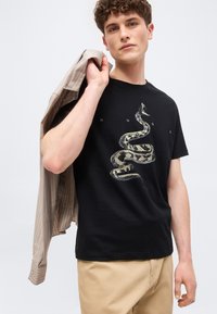 Νεαρός με σγουρά μαλλιά που φοράει μαύρο μπλουζάκι με μεγάλο γραφικό φίδι και μπεζ παντελόνι, κρατώντας ριγέ μπεζ πουκάμισο στον ώμο.