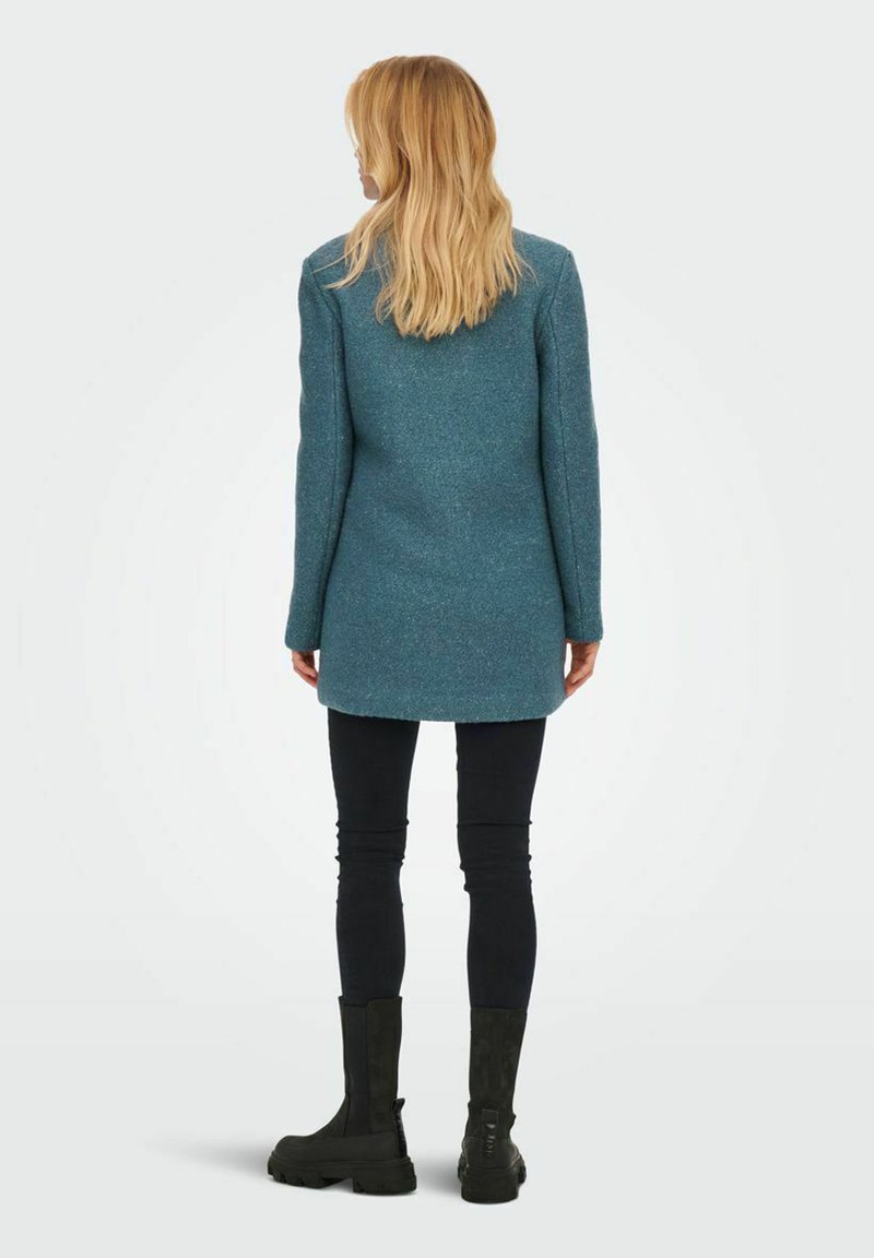 Türkisfarbene Wolljacke mit strukturiertem Finish, in mittellanger Länge, kombiniert mit schwarzen Skinny-Hosen und Ankle-Boots. Die Jacke hat einen geraden Schnitt und lange Ärmel.