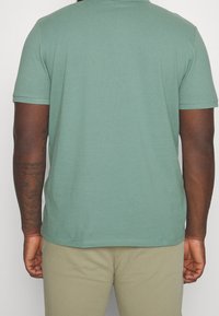 Polo shirt in cotone verde con colletto classico e maniche corte, caratterizzato da una texture liscia e orlo dritto. Indossato con pantaloni beige.