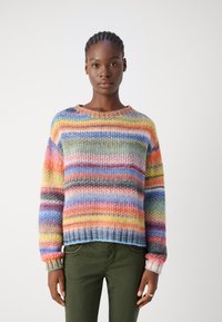 Vrouw met gevlochten haar die een meerkleurige gestreepte gebreide trui en olijfgroene broek draagt, tegen een witte achtergrond.