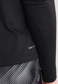 Mustat pitkähihiset urheiluvarusteet, jotka on valmistettu kosteutta siirtävästä kankaasta ja joissa on "DRI-FIT" -logo. Sileä rakenne, yhdistettynä kuvioituihin harmaisiin shortseihin.