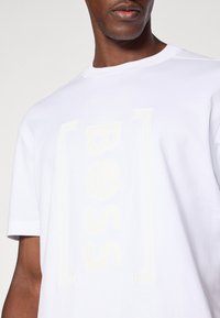 Λευκό βαμβακερό T-shirt με ανάγλυφο κρεμ λογότυπο "BOSS" και σχεδίαση ορθογωνίου περιγράμματος. Έχει κοντά μανίκια και στρογγυλή λαιμόκοψη.