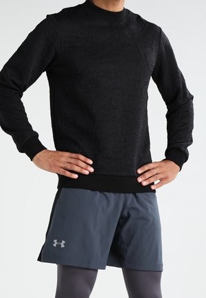 Mann trägt ein schwarzes, strukturiertes Langarmshirt, dunkelgraue Under Armour Shorts und graue Leggings, die Hände in die Hüften gestützt.