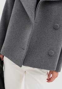 Grau wollener Mantel mit einem doppelt geknöpften Design, Reverskragen, texturiertem Stoff und großen runden Knöpfen. Weiße Hosen sind teilweise sichtbar.