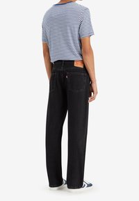 Personne portant un jean noir à jambes droites, un t-shirt à manches courtes rayé blanc et bleu, et des baskets blanches avec des accents bleus, vue de dos.