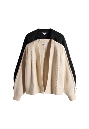 Kobieta w beżowym kardiganie i czarnym koronkowym crop topie stojąca ze skrzyżowanymi ramionami i uśmiechająca się na białym tle.