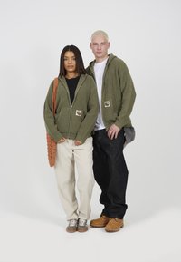 Green ribbed zip-up hoodies with patch details; one model in cream trousers and another in dark jeans, brown boots, and grey shoes.