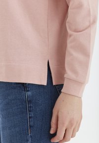 Top de algodón de manga larga en rosa claro con una abertura lateral en el dobladillo, combinado con jeans de mezclilla azul oscuro. El puño es ajustado y texturizado.