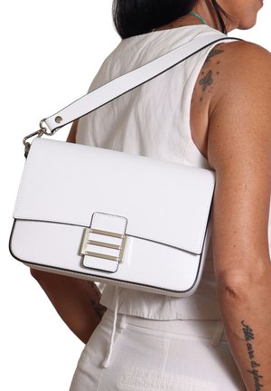 Sac en cuir blanc porté en bandoulière avec une boucle en argent, porté par une personne en haut sans manches blanc et pantalon, montrant des tatouages sur le bras et l'épaule.