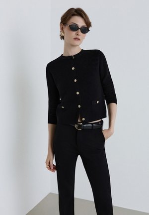 Frau mit kurzen Haaren, die schwarze Sonnenbrille, eine schwarze, geknöpfte Strickjacke, schwarze Hose und goldene Ohrringe trägt, steht vor einer schlichten hellen Wand.