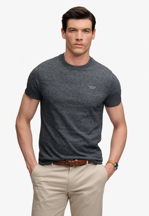 Hombre vistiendo una camiseta ajustada gris oscuro con un pequeño logo, pantalones beige, cinturón trenzado marrón y reloj de pulsera, de pie con las manos en los bolsillos.