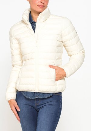 Donna che indossa un piumino bianco trapuntato con la zip chiusa sopra una camicia di jeans blu e dei jeans blu, con una mano in una tasca del giubbotto.