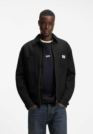Jeune homme portant une veste zippée noire sur un sweat Hugo noir et un jean bleu, debout les mains dans les poches devant un fond blanc.