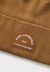Hnědá pletená čepice s texturovaným povrchem a koženým štítkem s nápisem "KARL LAGERFELD PARIS" a adresou embosovanou bílou barvou.