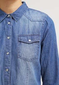 Camicia in denim di colore blu chiaro con colletto classico, bottoni a pressione e taschino sul petto con pattina e chiusura a pressione. Tessuto morbido e strutturato.