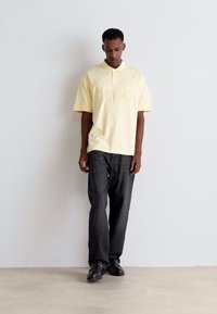 Mann, der gegen eine weiße Wand steht, trägt ein blassgelbes Poloshirt und lose dunkelgraue Hosen mit schwarzen Schuhen.