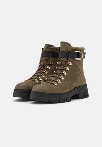 Stivali da trekking in suede verde oliva con una fascia nera, occhielli dorati e una suola nera spessa. Presentano un dettaglio con fibbia alla caviglia.