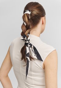 Foulard en soie noué autour d'une coiffure tressée. Le foulard présente un motif géométrique noir et blanc avec de longues extrémités flottantes. Haut neutre.