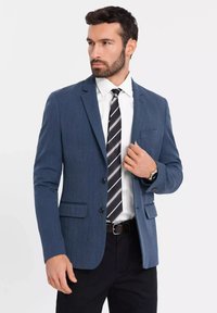 Niebieska marynarka o teksturowym wykończeniu, z dwiema przednimi kieszeniami, zapięciem na jeden guzik, połączona z białą koszulą i paskiem w stripes.