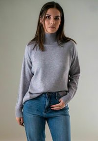 Jonge vrouw met lang bruin haar, gekleed in een lichtgrijze coltrui en blauwe spijkerbroek, staande voor een effen grijze achtergrond.