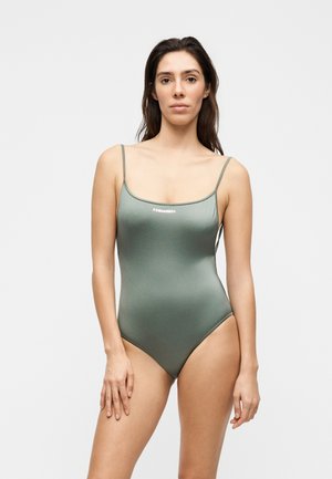 Γυναίκα που στέκεται, φοράει μια εφαρμοστή γκρι-πράσινη ολόσωμη φόρμα με λεπτές τιράντες και μικρό λευκό λογότυπο "DSQUARED2" στο στήθος.