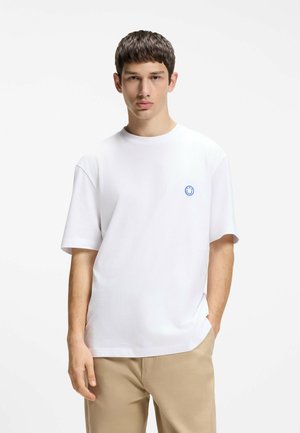 Joven con una camiseta blanca lisa con un pequeño logotipo azul de una cara sonriente y pantalones beige, de pie con una mano en el bolsillo.