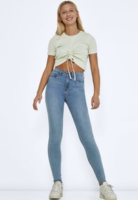 Top corto de canalé verde claro con fruncido frontal y detalle de lazo, combinado con jeans ajustados de color azul claro y zapatillas blancas. Fondo liso.