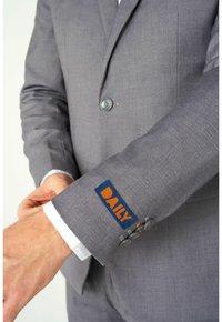 Grau Anzugjacke mit einem dekorativen dunkelblauen Etikett mit "DAILY" in Orange am Ärmel, akzentuiert mit zwei grauen Knöpfen.