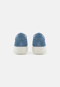 Baskets en denim avec une tige bleue, une semelle en caoutchouc blanc et une texture douce. Les caractéristiques incluent des accents de couture et des langues de talon en boucle pour un enfilage facile.