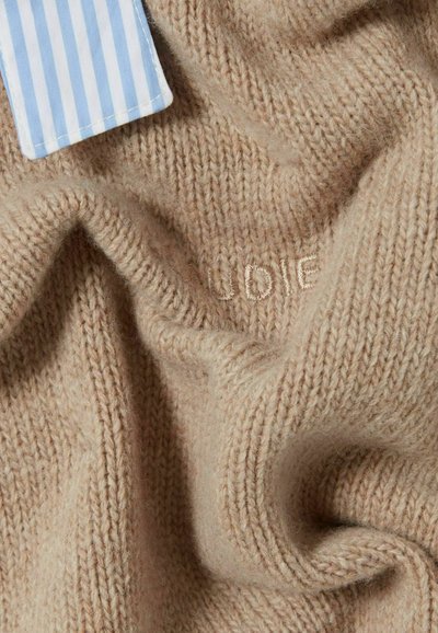 Tessuto lavorato a maglia beige con una morbida texture, caratterizzato da cuciture sottili che formano la scritta "UDIE" e un'etichetta a righe blu e bianche attaccata.