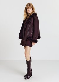 Giacca in montone bordeaux con zip frontale, colletto peloso e polsini pelosi. Abbinata a un mini vestito e stivali lucidi alti fino al ginocchio.