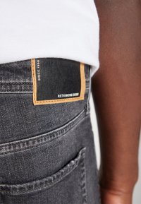 Mörkgrå denimjeans med ett läderpatch som har texten "RETHINKING DENIM" och en beige detalj, synlig sömnad och detaljer på bakfickan.