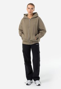 Zip-up hoodie in olive green, with a relaxed fit, front pockets, and ribbed cuffs. Paired with black cargo pants and white sneakers.