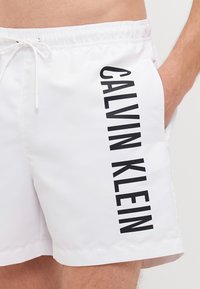 Biele plavky vyrobené z hladkej tkaniny s tlačeným čiernym logom "CALVIN KLEIN" vertikali pozdĺž strany. Nastaviteľný pás s šnúrkou.