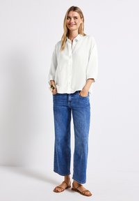Blusa blanca de botones con un corte holgado y mangas tres cuartos, combinada con jeans amplios azules y sandalias planas marrones.