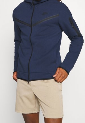 Mann trägt eine marineblaue Nike Jacke mit Reißverschluss und schwarzen Akzenten sowie beige Shorts, steht mit einer Hand in der Tasche.
