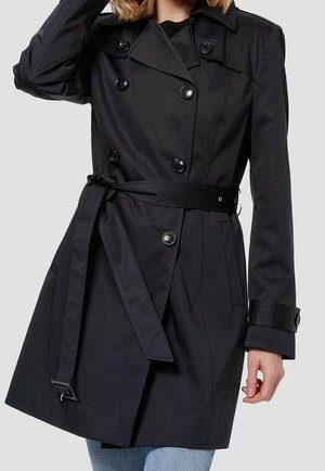 Nainen pukeutuneena istuvaan mustaan kaksiriviseen trenssitakkiin, jonka vyö on löysästi sidottu, ja sinisiin farkkuihin, käsi nostettuna pään lähelle.