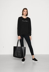 Felpa nera con collo rotondo e ricamo "Tommy Hilfiger", pantaloni neri, borsa tote nera e sneakers nere con suole bianche.