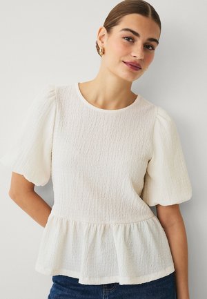 Ung kvinde iført en struktureret hvid bluse med puffede ærmer og peplumkant, kombineret med mørkeblå jeans, foran en ensartet lys baggrund.