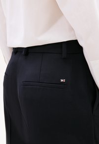 Marineblauwe broek van gestructureerde stof met een slim fit, voorzien van een achterzak en een klein metalen logoaccent. Gecombineerd met een lichtgekleurde blouse.