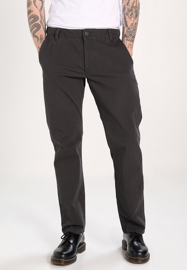 Homme portant un pantalon noir droit, des chaussures noires à lacets, et une chemise blanche avec les avant-bras tatoués visibles, debout les mains dans les poches.