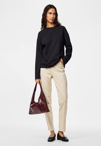 Donna con capelli lunghi e ricci che indossa una maglia nera a maniche lunghe, pantaloni beige, scarpe nere, tenendo una borsa di pelle rossa scura.