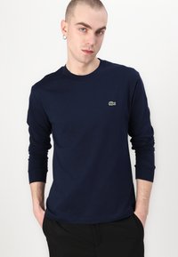 T-shirt à manches longues bleu marine en tissu coton, avec un col rond et un petit logo crocodile vert sur la poitrine gauche. Texture lisse.