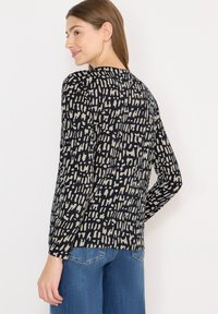 Kvinde iført en langærmet sort og beige mønstret bluse og blå jeans, set fra siden med et let smil på en hvid baggrund.